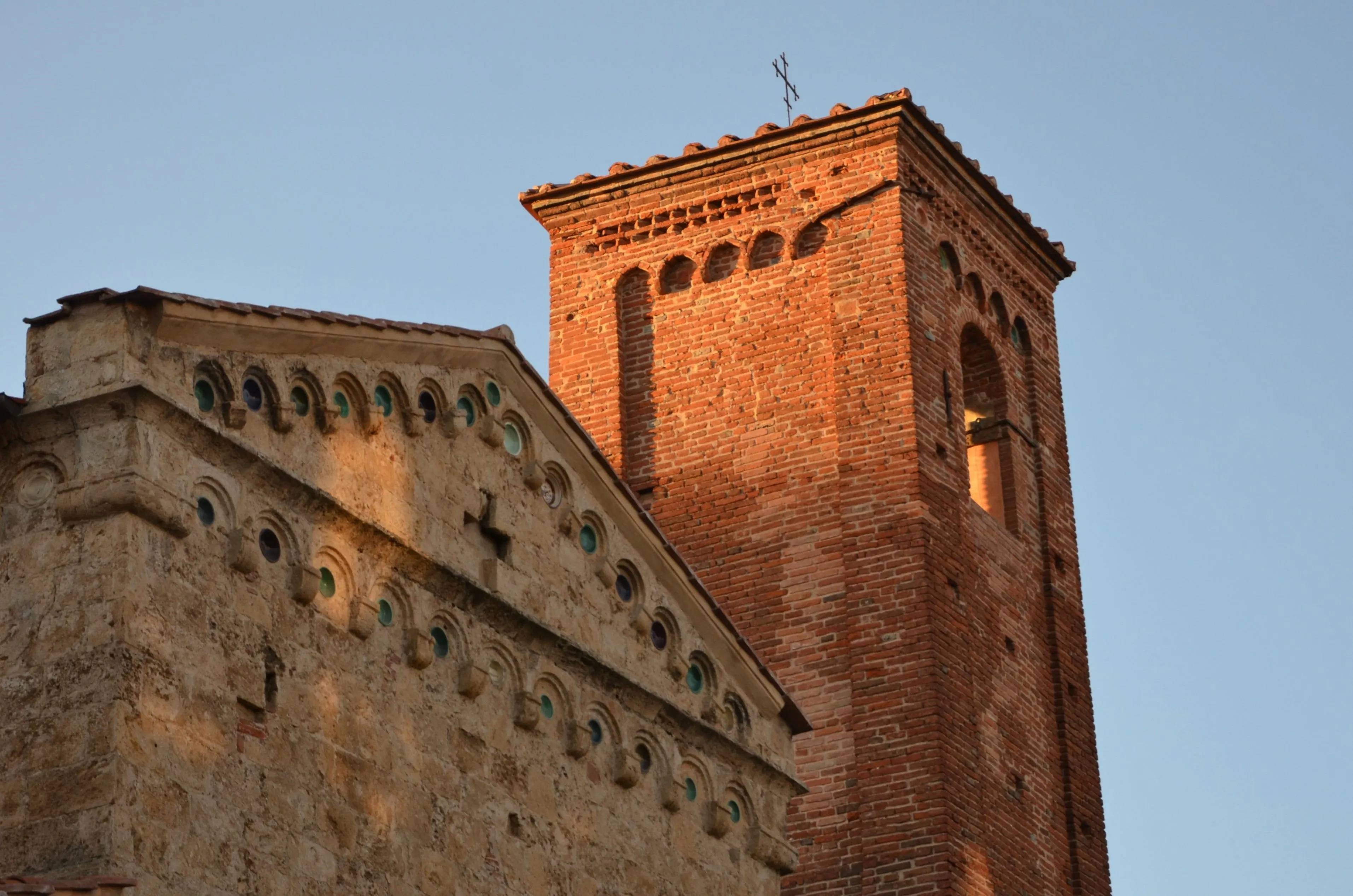 A close-up, editorial-style image of an ancient brick church tower and an adjacent stone facade with intricate decorative elements, illuminated by warm, directional sunlight, set against a clear blue sky.