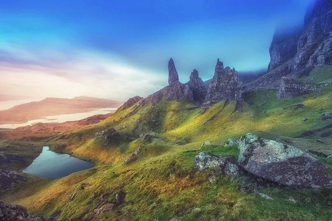 A dramatic landscape featuring prominent rock formations (possibly the Old Man of Storr), rolling green hills, and a tranquil body of water under a vibrant, clear blue sky.