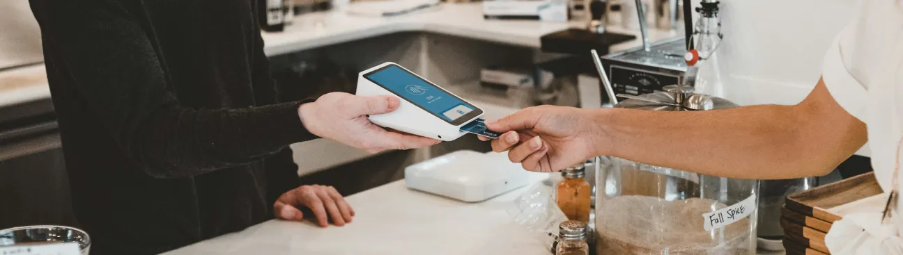 An editorial image capturing a credit card transaction at a point-of-sale terminal. Two hands are visible, one holding the payment device and the other inserting a credit card. The primary subjects are in sharp focus, while the background is subtly blurred to emphasize the action.
