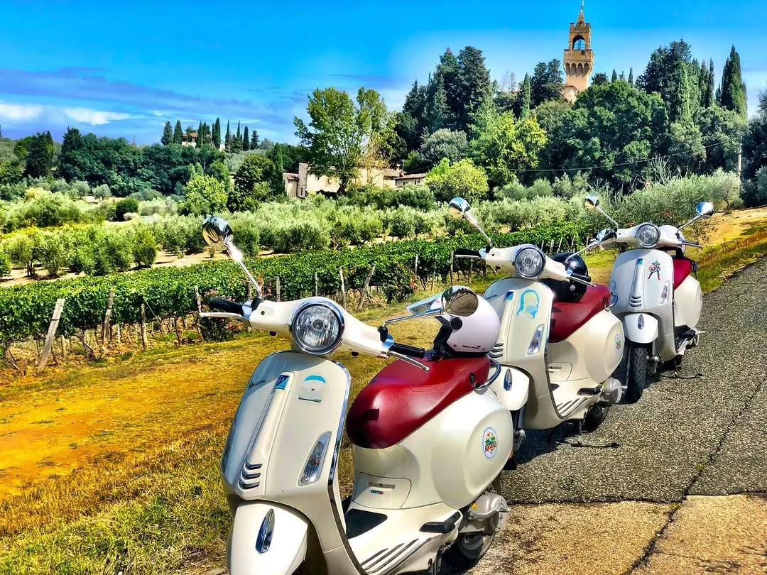A picturesque view of a row of white and red Vespas parked on a paved path, overlooking a vibrant Tuscan landscape with vineyards, olive groves, and a distant historic tower under a clear blue sky.