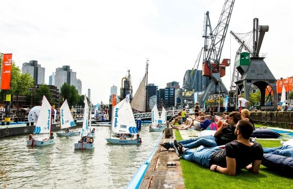 The image successfully captures a vibrant urban scene featuring small sailboats navigating a waterway, with people relaxing on the grassy bank and industrial cranes and city buildings in the background. It effectively conveys the atmosphere of a community event or leisure activity in an urban setting.