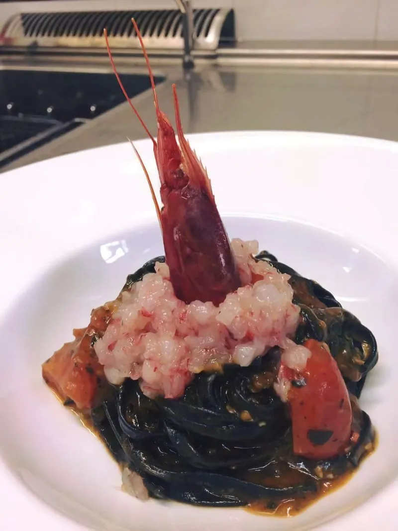 A close-up shot of a gourmet dish featuring squid ink pasta topped with a large, cooked red prawn and a portion of what appears to be prawn tartare, served in a white ceramic bowl.
