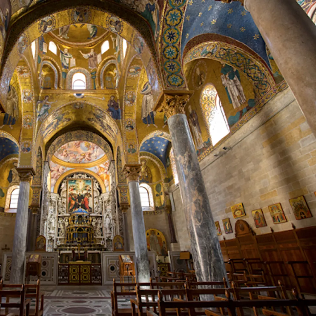An interior shot of an ornate church or cathedral, featuring intricate gold mosaics covering the vaulted ceilings and walls. Large marble pillars frame the central view towards the altar. The lighting appears natural, highlighting the rich details of the space.