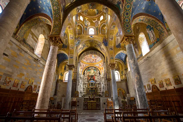 An interior shot of an ornate church or cathedral, featuring intricate gold mosaics covering the vaulted ceilings and walls. Large marble pillars frame the central view towards the altar. The lighting appears natural, highlighting the rich details of the space.