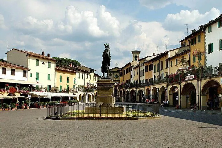 A sunny wide shot of a European town square with a large statue in the center, surrounded by historic buildings with arches and green window shutters. The sky is bright with scattered clouds.