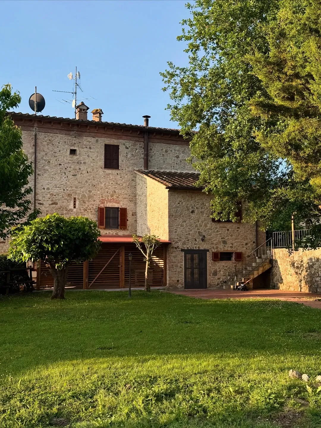 A stone-built house with a red-tiled roof and brown shutters, surrounded by green grass and trees, under a clear blue sky. The sunlight casts shadows, highlighting the textured stone facade.