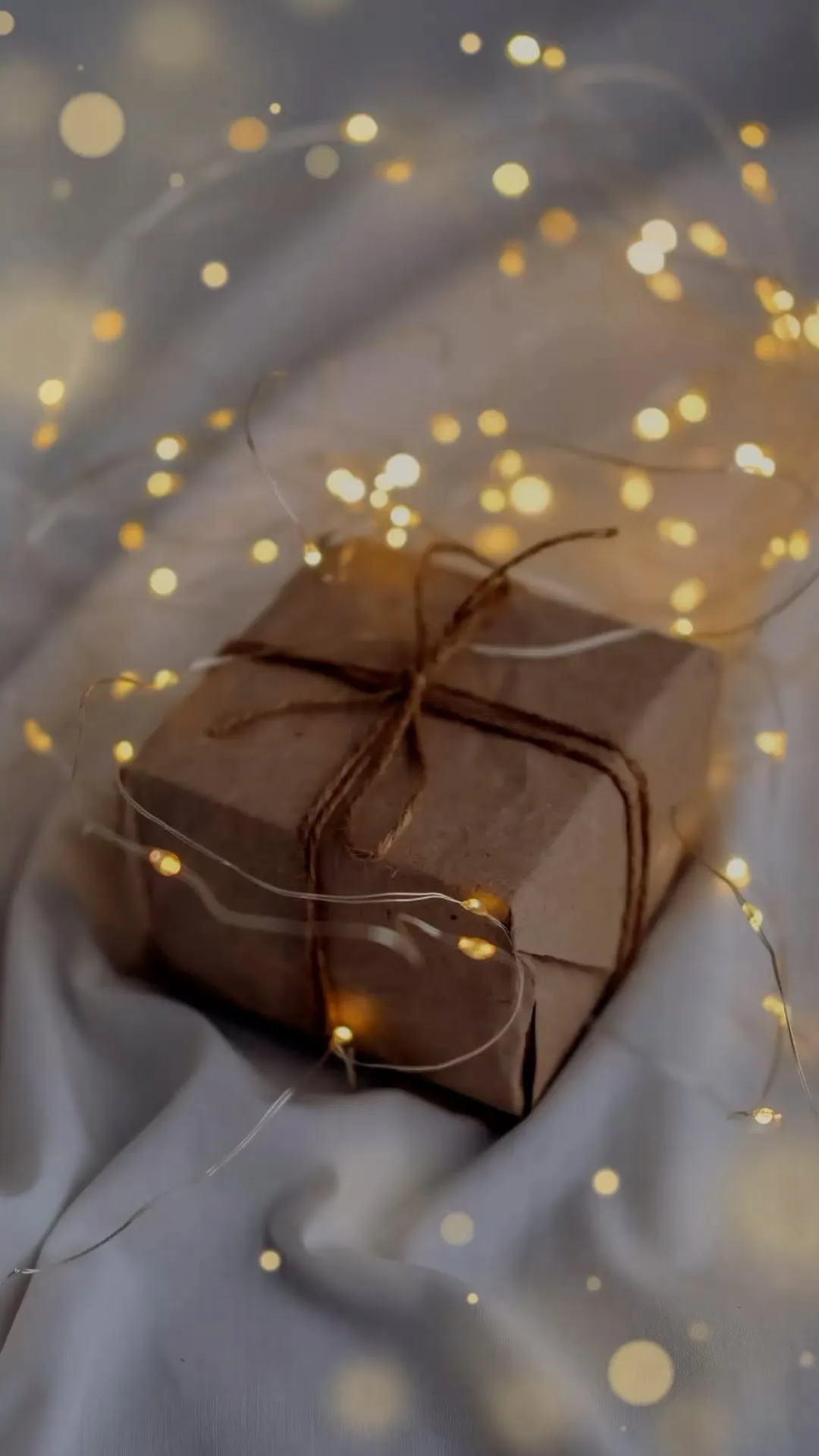 A warm and atmospheric image of a brown paper-wrapped gift tied with twine, surrounded by glowing fairy lights, resting on a soft, light-colored fabric.