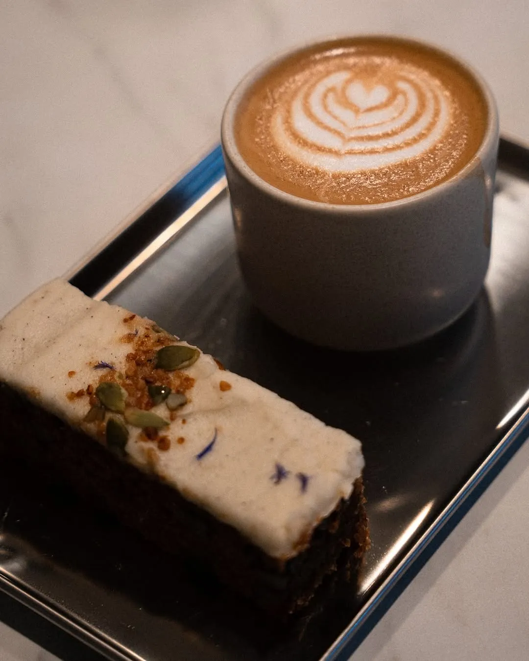 A close-up shot of a slice of cake topped with white frosting, pumpkin seeds, and small flower petals, positioned next to a cup of coffee with intricate latte art. Both items are placed on a reflective metallic tray, hinting at a cafe or dessert setting.
