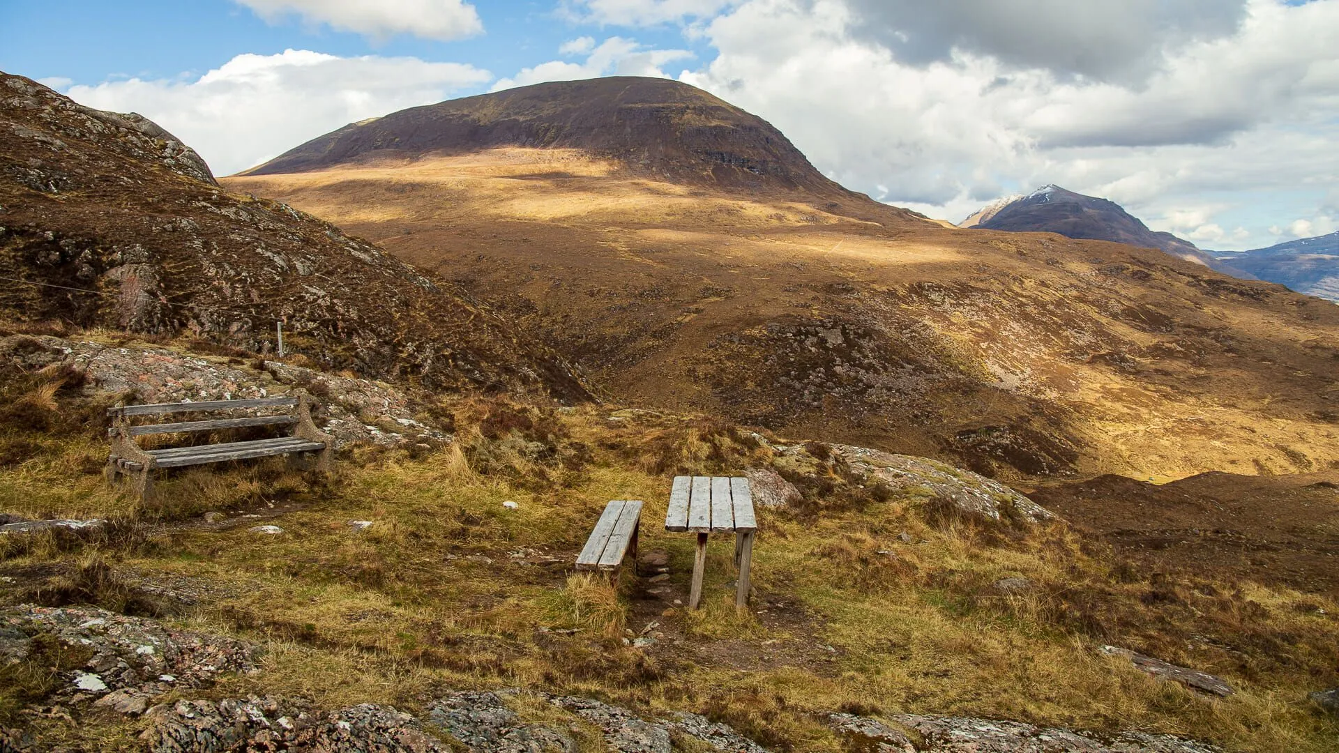 The image features a well-composed landscape with a wooden picnic table and bench in the foreground, leading the eye towards a large, prominent mountain under a partly cloudy sky. The lighting is natural and highlights the textures of the terrain. The image is sharp throughout, with both foreground and background elements in clear focus. Colors are accurate, reflecting the earthy tones of the mountainous region. The photograph is highly photo-realistic.