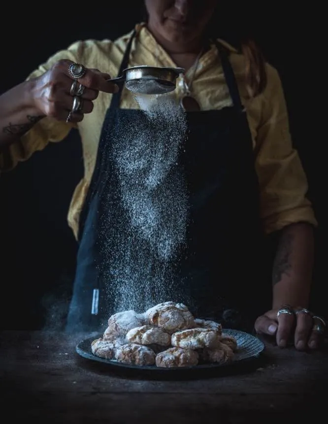 The image is a well-composed editorial-style shot capturing the action of sifting powdered sugar over cookies. The dramatic and focused lighting effectively highlights the falling sugar and the textured cookies, creating a strong visual impact. The image is sharp, with all key elements in focus, contributing to its high photo-realistic quality and overall aesthetic.