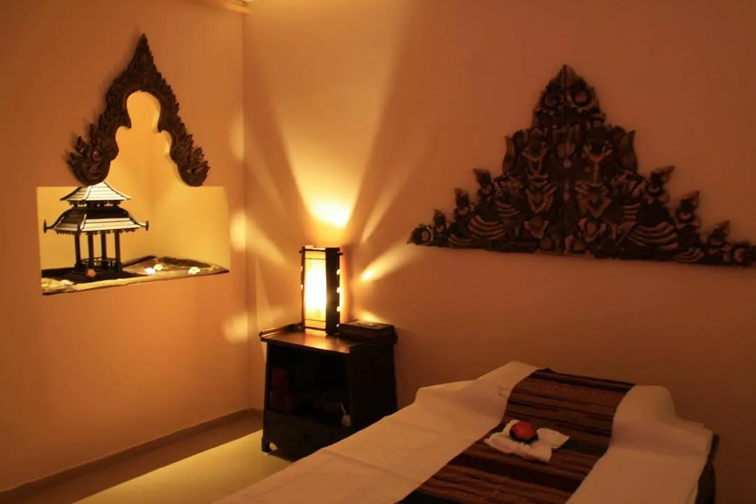 An interior shot of a serene spa or massage room, featuring a massage bed, a wooden side table with a lamp casting warm, atmospheric light, and traditional decorative elements on the walls.