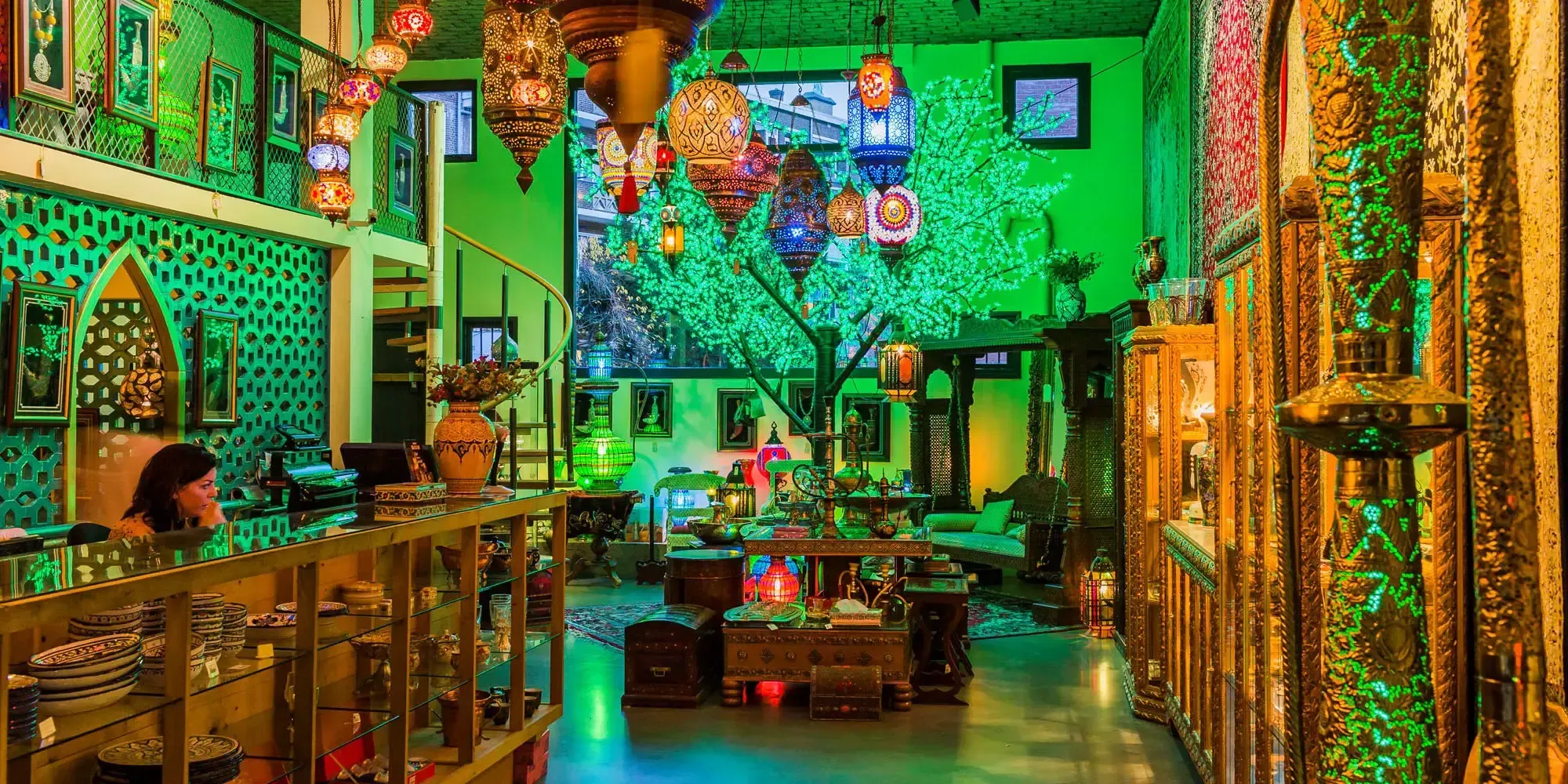 An image showcasing a vibrant and richly decorated interior space, likely a shop or gallery, filled with an array of ornate, colorful lanterns hanging from the ceiling. A prominent tree-like structure with bright green illumination stands centrally, casting a strong emerald glow across the scene. The room also features intricate furniture, decorative items, and a spiral staircase on the left, all contributing to a maximalist and exotic aesthetic.