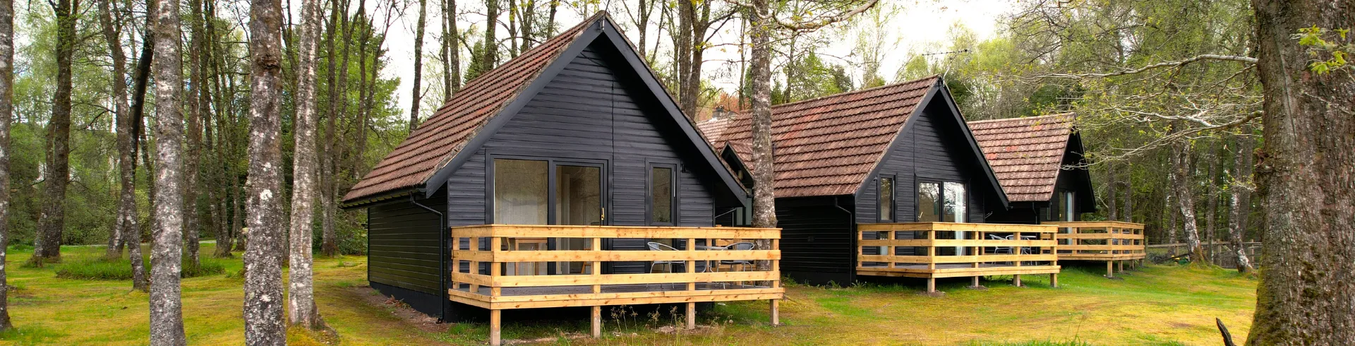 An exterior shot of modern black cabins with traditional tiled roofs, nestled among trees. A wooden deck and railing are visible in the foreground, with outdoor furniture on the deck.