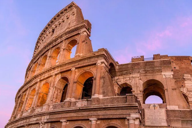 The image features a close-up, upward-angled shot of the Colosseum in Rome, capturing its imposing ancient architecture against a soft blue and pink sky. The composition is strong, emphasizing the scale and detail of the structure, particularly its arches and stone textures. The lighting is natural and even, highlighting the building without harsh shadows. The image is sharp and photo-realistic, with accurate and pleasing colors.