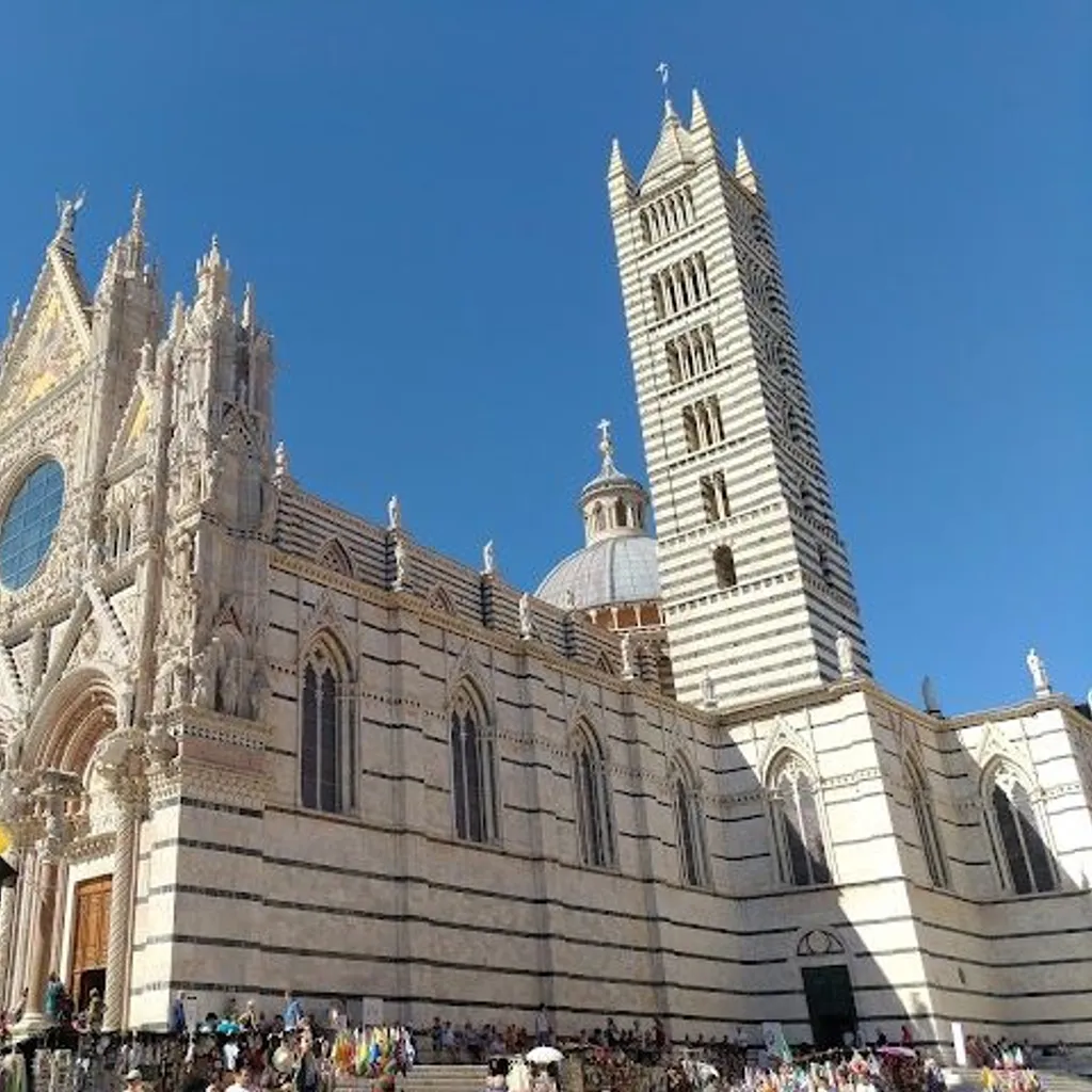 A grand, intricately detailed cathedral, possibly the Siena Duomo, stands against a clear, vibrant blue sky on a sunny day. The lower portion shows a crowd of people gathered around the base of the building, with some colorful flags visible.
