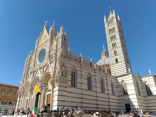 A grand, intricately detailed cathedral, possibly the Siena Duomo, stands against a clear, vibrant blue sky on a sunny day. The lower portion shows a crowd of people gathered around the base of the building, with some colorful flags visible.