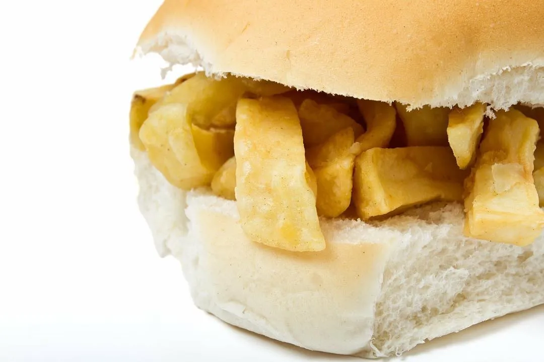 A close-up shot of a chip butty, featuring golden-brown chips nestled within a soft white bread bun, presented against a clean white background.