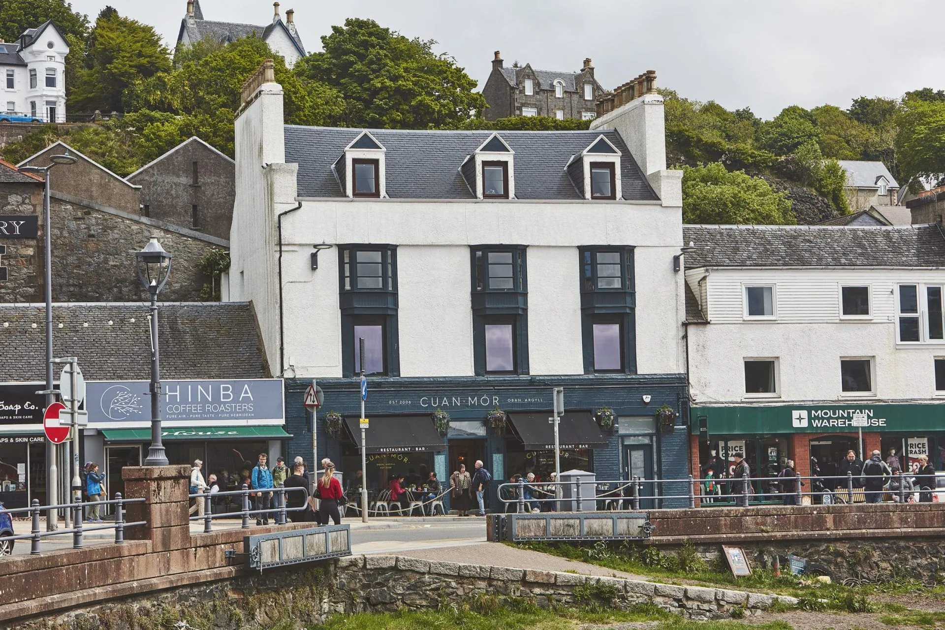 An exterior view of a white and dark blue building, likely a restaurant or bar, situated on a street with pedestrians. The building has multiple windows and dormer roofs, with a stone bridge and other buildings visible in the background under an overcast sky.