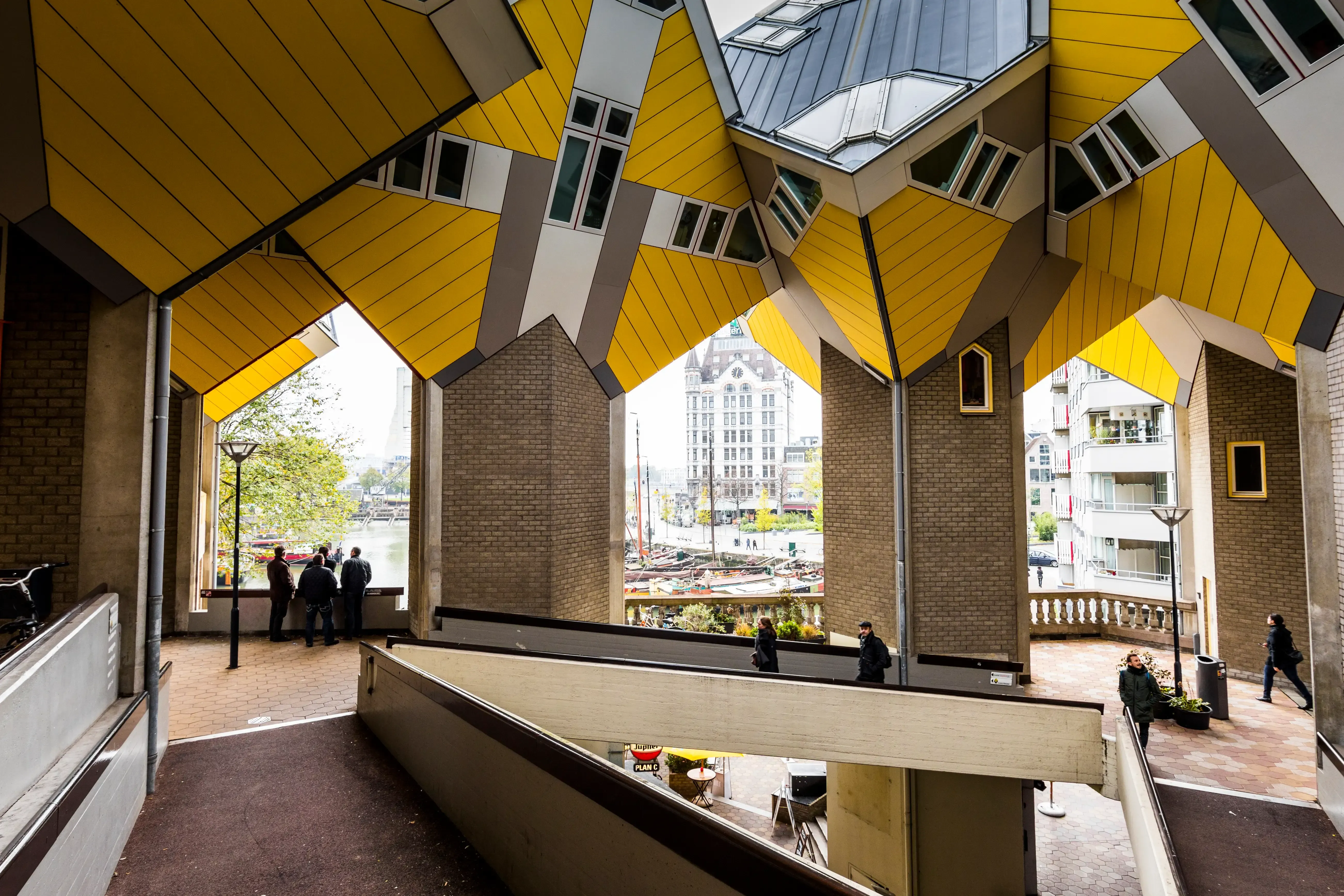 This image brilliantly captures the distinctive architecture of the Cube Houses in Rotterdam, using their geometric forms to frame a dynamic view of the city's canal and distant buildings. The composition is well-balanced and uses leading lines effectively. The lighting is natural and even, ensuring clarity and detail throughout the scene. Sharpness is excellent, with fine details visible in both the foreground architecture and the background elements. Colors are accurate and vibrant, particularly the iconic yellow of the buildings. The image is highly photo-realistic, and all key elements are in sharp focus, making it a compelling editorial-style architectural photograph.