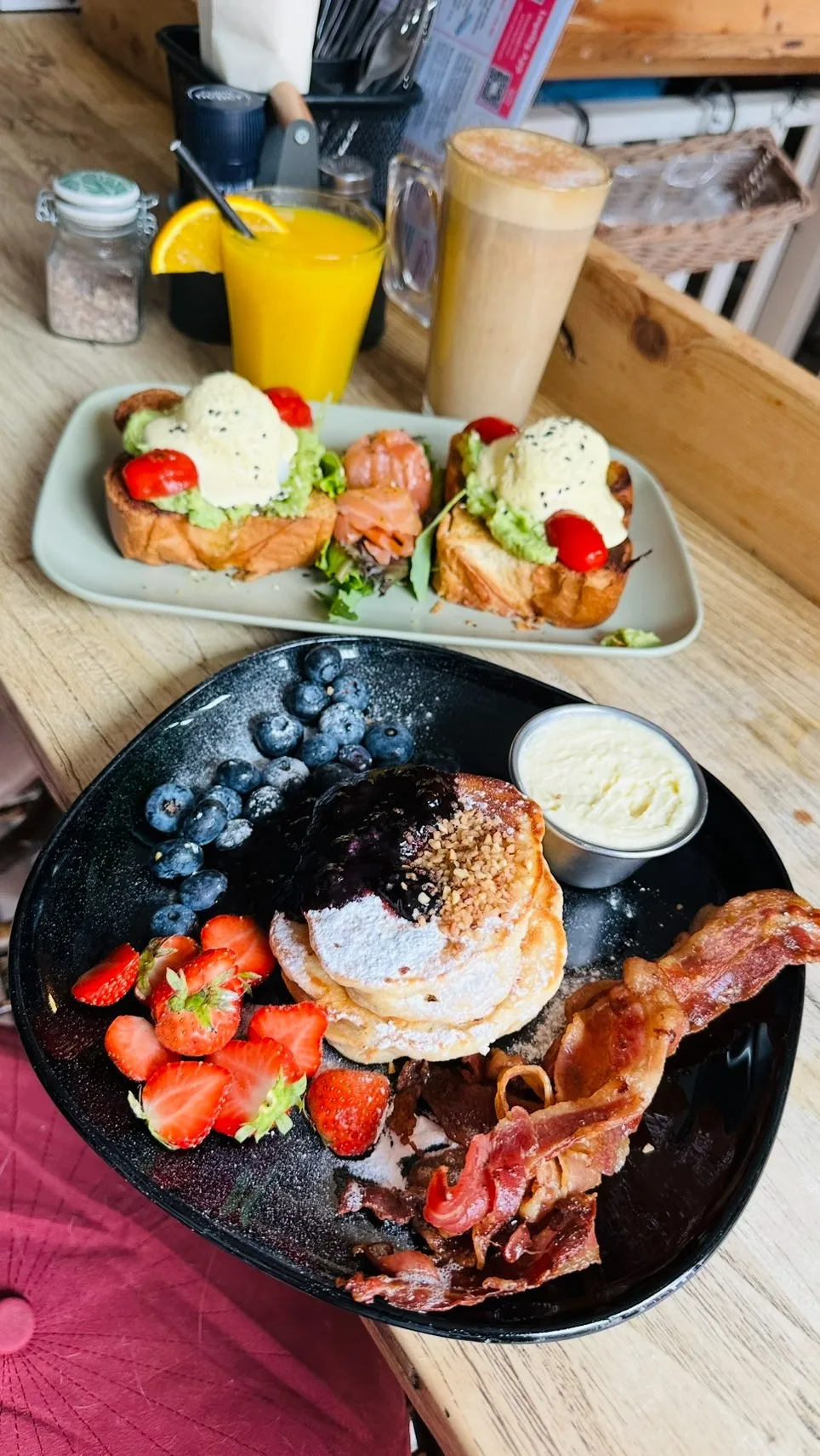 A vibrant image displaying two breakfast plates on a wooden table. The foreground plate features pancakes with blueberries, strawberries, bacon, and a side of cream. The background plate holds two avocado toasts topped with poached eggs and cherry tomatoes, alongside smoked salmon.