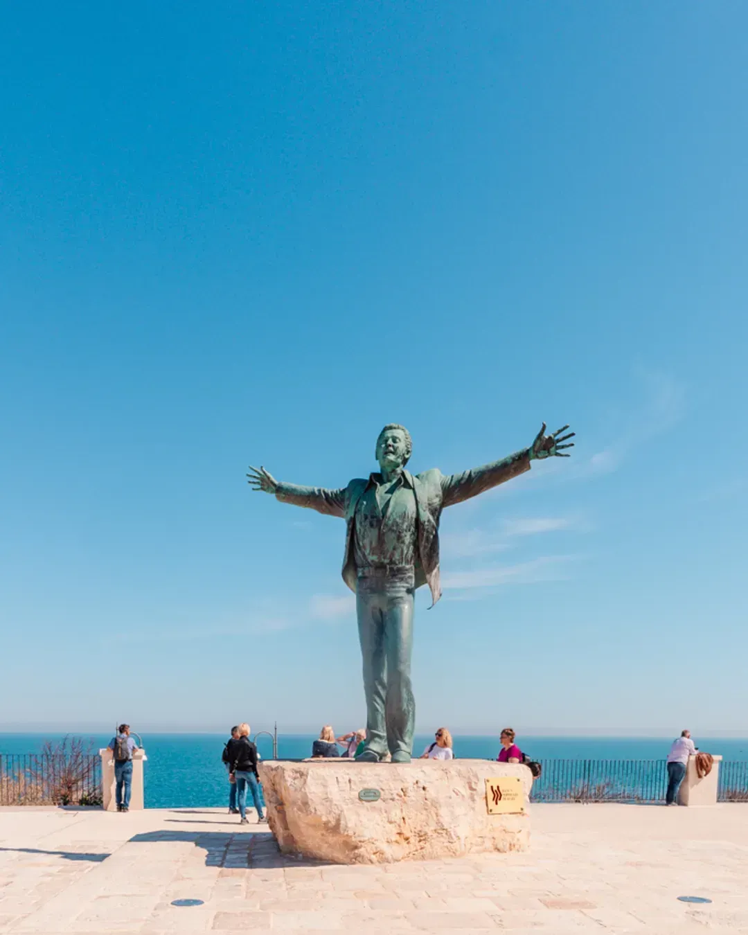 An editorial image of a bronze statue of Domenico Modugno in Polignano a Mare, Italy, with arms outstretched, set against a vast, clear blue sky and the Adriatic Sea.