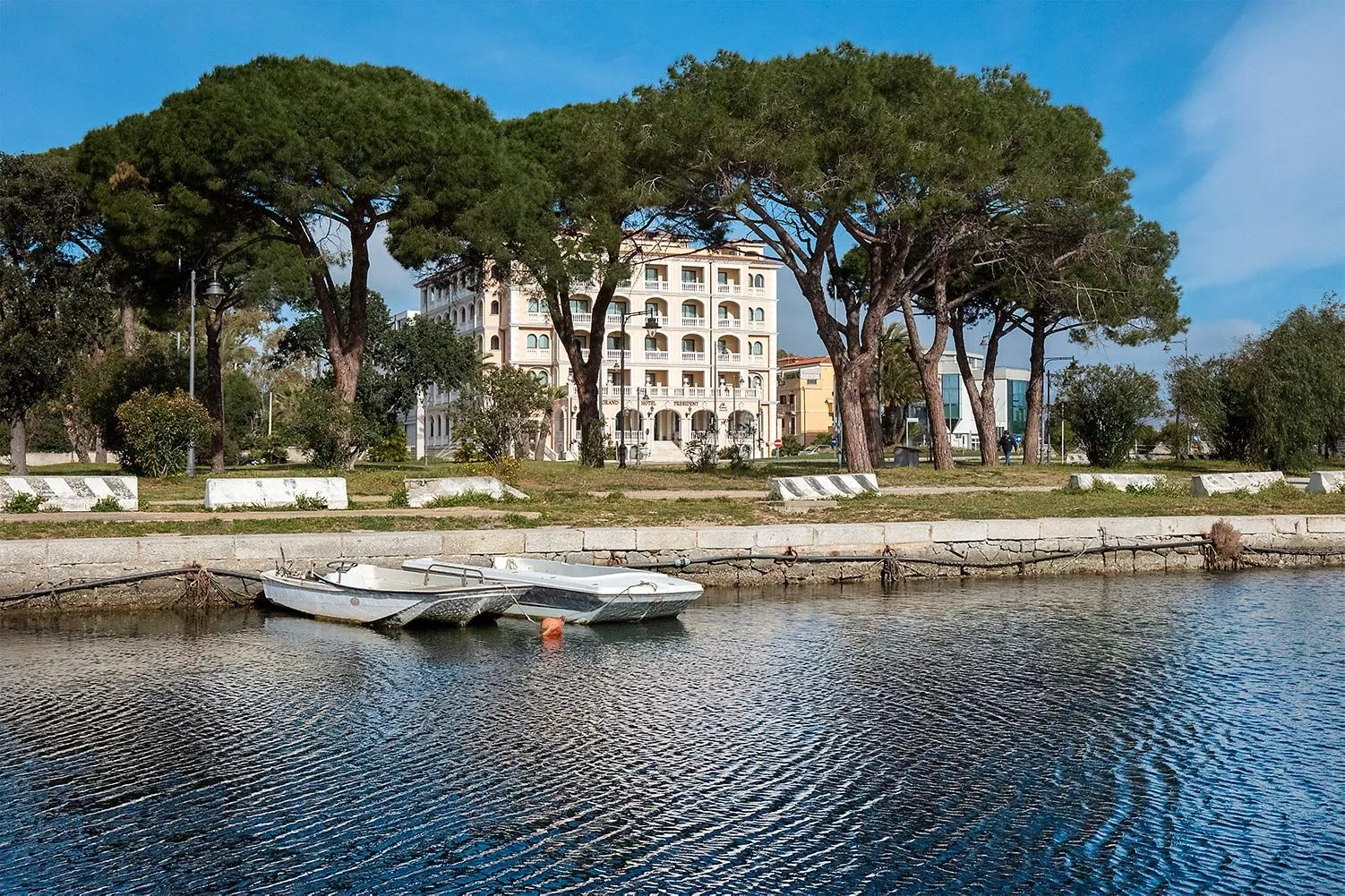 An editorial-style image showcasing a multi-story building with architectural details, framed by large trees and a body of water with two small boats in the foreground. The scene appears to be set on a clear, sunny day.