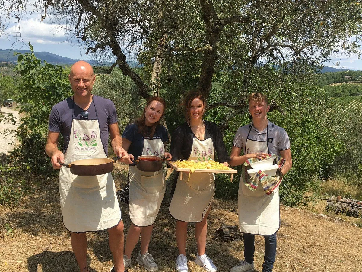 A group of four people, likely family or friends, wearing aprons and holding cooking utensils and ingredients, standing outdoors under a tree, possibly during a cooking class or culinary experience. They appear to be making pasta, with one person holding a tray of raw pasta shapes and another holding a pot with sauce.