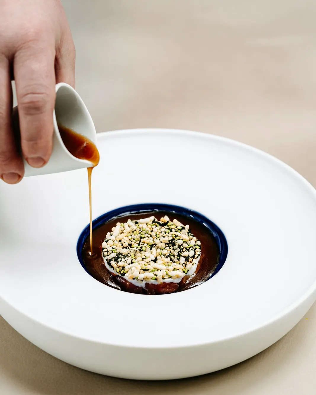 A close-up, high-angle shot of a hand pouring a rich brown sauce from a white pitcher onto a meticulously prepared dish in a modern white bowl with a dark blue inner rim. The dish features a base topped with white and green crunchy elements, suggesting a gourmet dessert or main course.