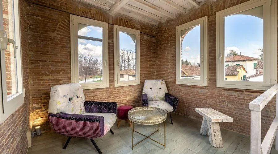 A cozy room featuring exposed brick walls, two decorative armchairs, a round coffee table, and three windows overlooking an outdoor scene. The room is well-furnished and appears inviting.