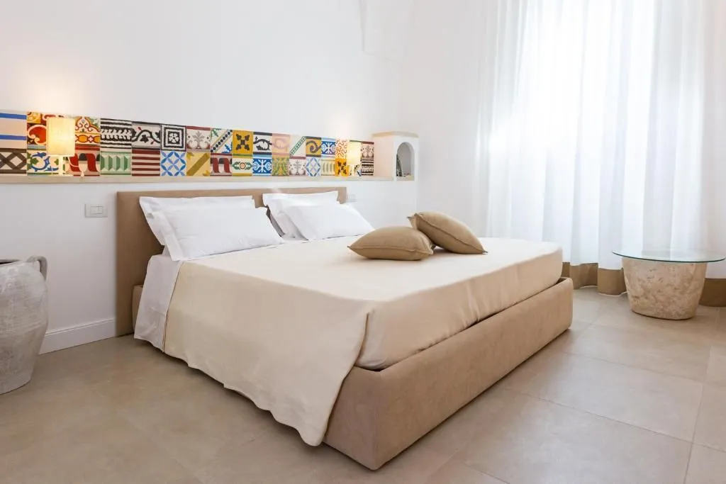 An image of a clean, minimalist bedroom featuring a bed with white and beige linens, a colorful tiled headboard, and bright natural light coming from a window with sheer curtains.