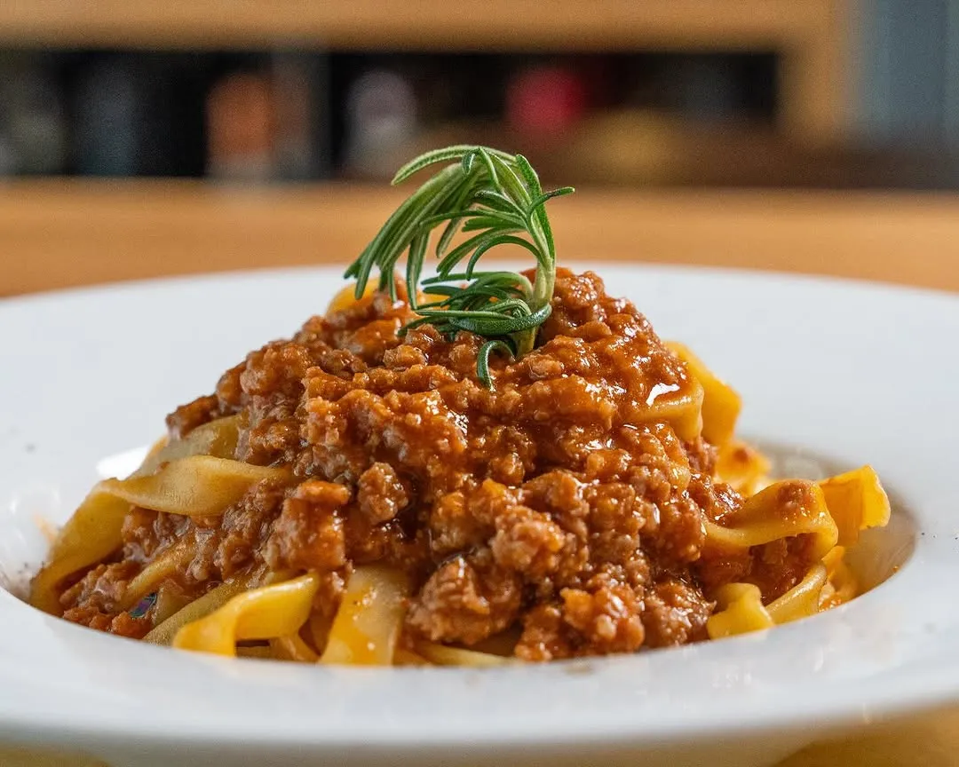 A well-composed and appetizing close-up of pasta with bolognese sauce, garnished with rosemary. The image is well-lit, sharp, and highly photo-realistic, showcasing the dish's texture and color accurately.