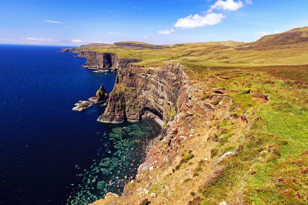 A stunning high-angle view of rugged coastal cliffs meeting the clear blue sea, with expansive green and brown grasslands stretching inland under a bright blue sky with sparse clouds.