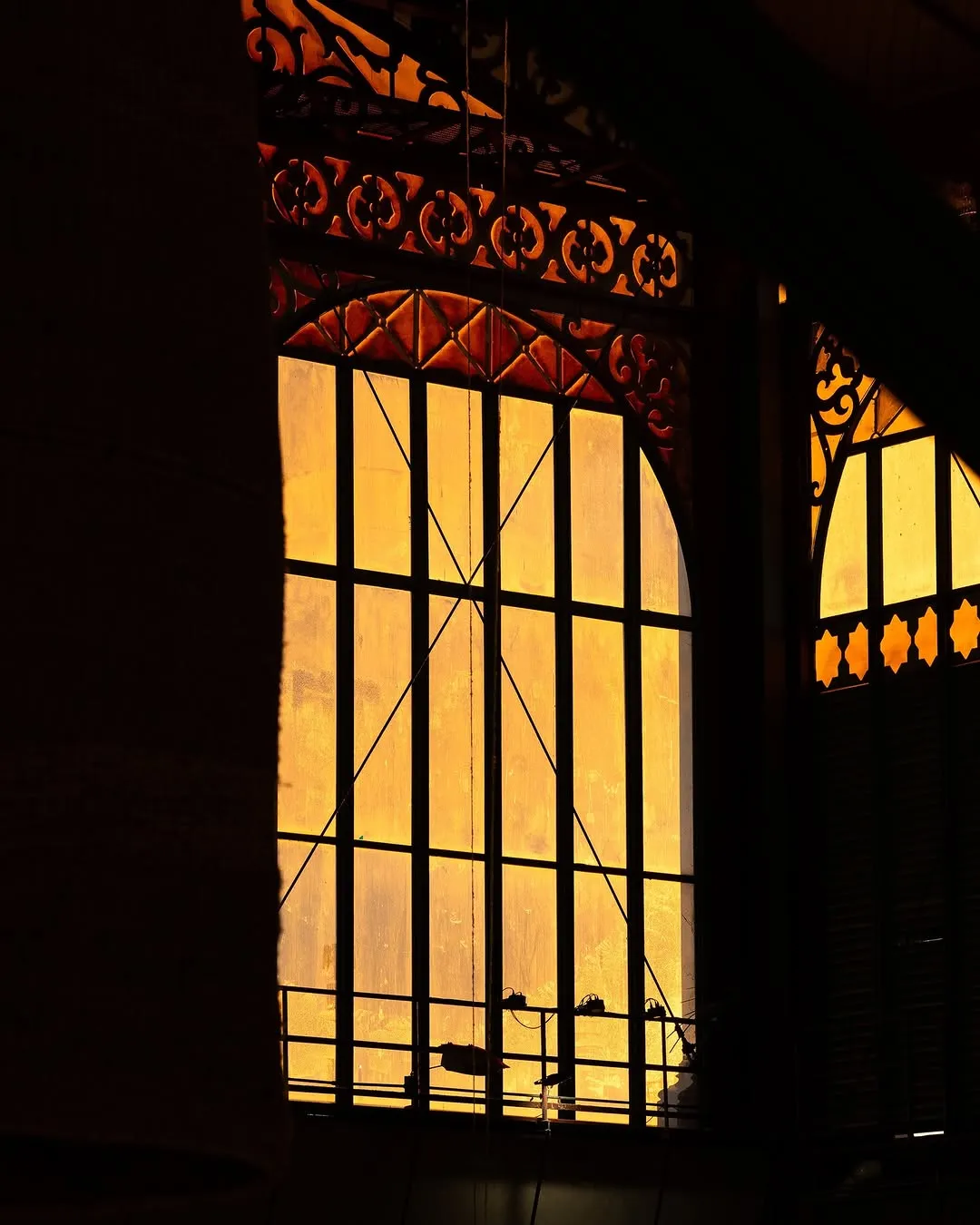 The image features a striking architectural detail of an ornate window, silhouetted against a vibrant golden light. The intricate metalwork of the window frame and its decorative elements are highlighted by the strong backlight, creating a high-contrast and dramatic composition. The surrounding interior is deeply shadowed, emphasizing the luminous quality of the window.