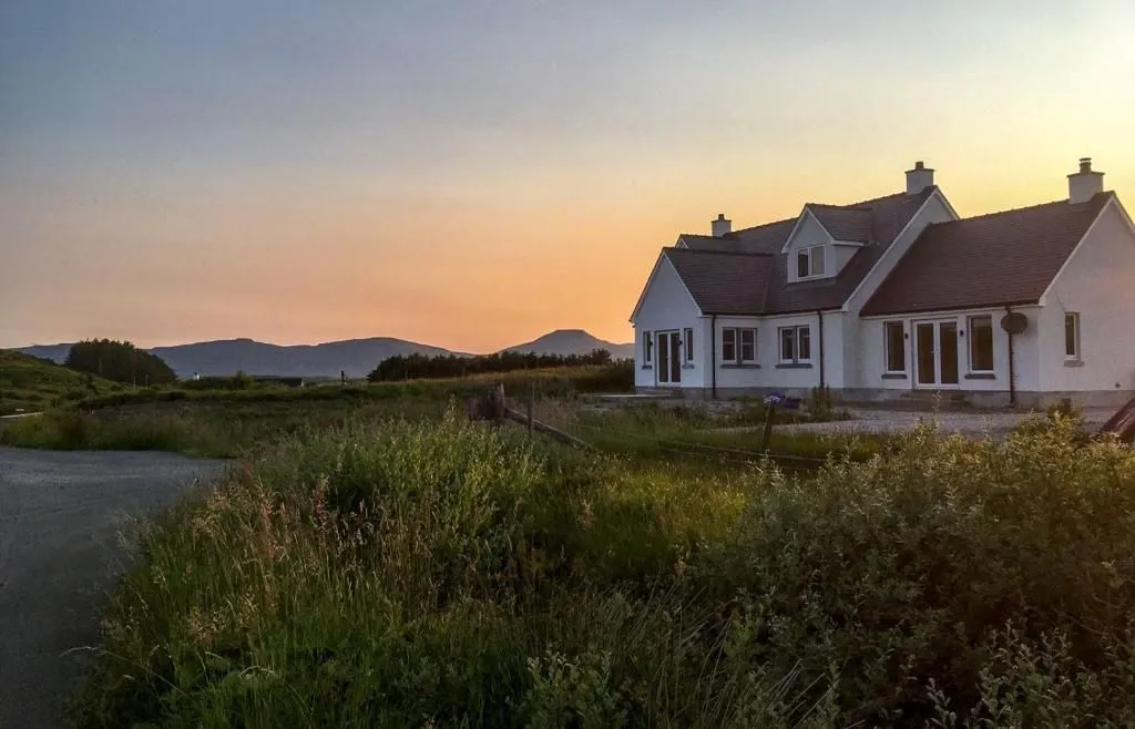 An image depicting a white house situated in a grassy field with mountains in the background under a sunset sky. The golden hour lighting casts warm tones across the scene.