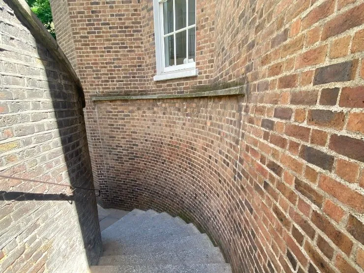 An exterior view of a curving brick wall with a white-framed window and a descending stone staircase. The image captures the architectural details under direct sunlight.