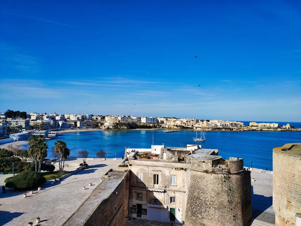 A high-angle view of a coastal town, featuring an old stone fortress in the foreground, a vibrant blue bay with a sailboat, and the town's buildings lining the coastline under a clear sky.