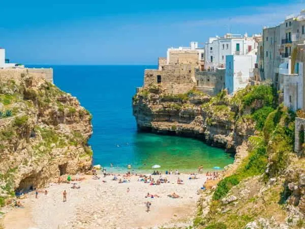 A beautiful, vibrant aerial shot of a Mediterranean beach cove, featuring clear turquoise waters, sandy shores with people, and ancient white buildings perched atop rugged cliffs under a bright blue sky.