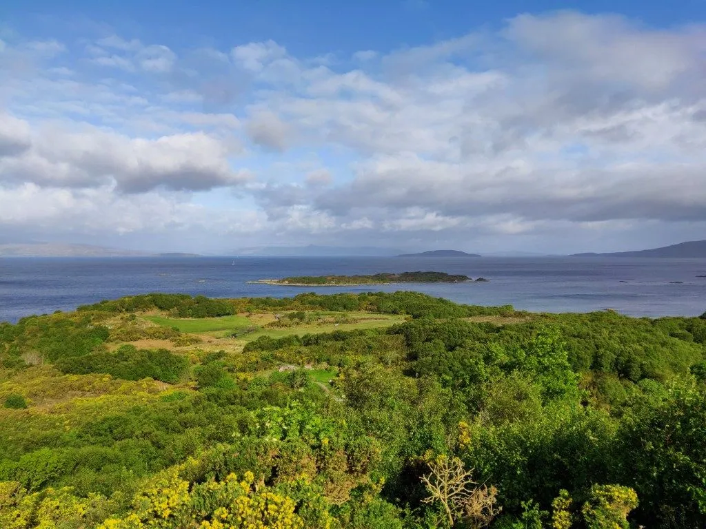 A vibrant and well-composed landscape image showcasing a coastal view with lush green foliage in the foreground, leading to a expansive blue sea dotted with distant islands, all under a bright sky with scattered clouds.