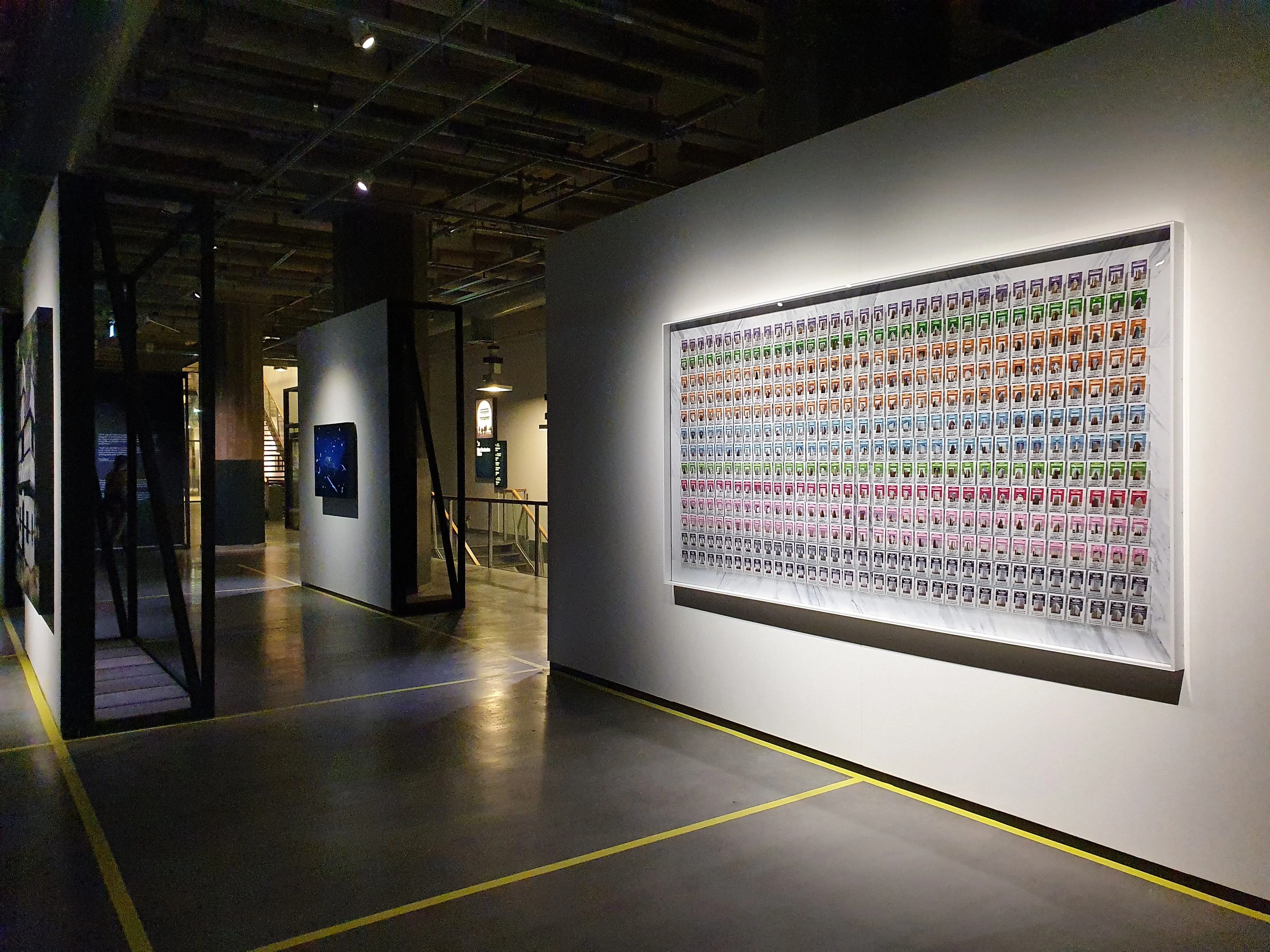 The image features an interior view of a museum or gallery, highlighting a large, brightly lit art display on a white wall. The composition effectively draws attention to the artwork, which appears to be a grid of colorful, small graphics or symbols. While the main subject is well-lit and sharp, the overall lighting in the surrounding space is dim, leading to some underexposed areas, particularly the ceiling and distant parts of the room. The image is photo-realistic, with accurate colors for the illuminated display, and key elements are in focus.
