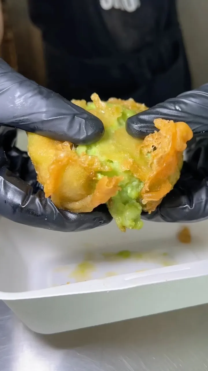 A close-up image showcasing a deep-fried food item, possibly an avocado fry or similar, being gently pulled apart by gloved hands to reveal its vibrant green interior and crispy golden-brown exterior.