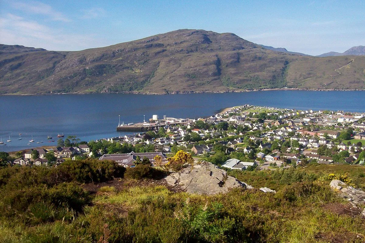 A panoramic shot capturing a scenic coastal town situated along a bay, with a large mountain serving as a prominent backdrop under a clear sky.