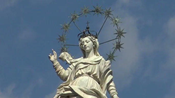 A well-composed photograph of a white marble statue, possibly a religious figure such as the Virgin Mary, adorned with a crown and a halo of stars, set against a clear blue sky.