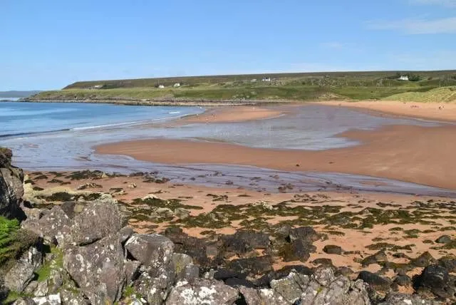 A bright, clear day illuminates a sandy beach at low tide, with wet sand reflecting the light. Large, dark rocks covered in some green moss dominate the foreground, leading the eye towards the calm blue water and the distant green hills under a clear sky.