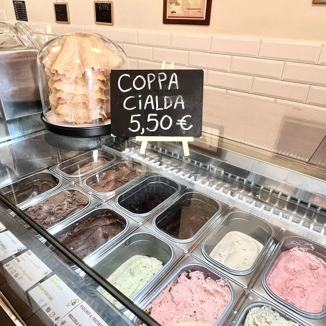 An eye-level shot capturing a vibrant display of various gelato flavors in a refrigerated counter, with a stack of waffle cones under a cloche and a small blackboard sign indicating 'COPPA CIALDA 5,50 €' visible above. The background features a white tiled wall.