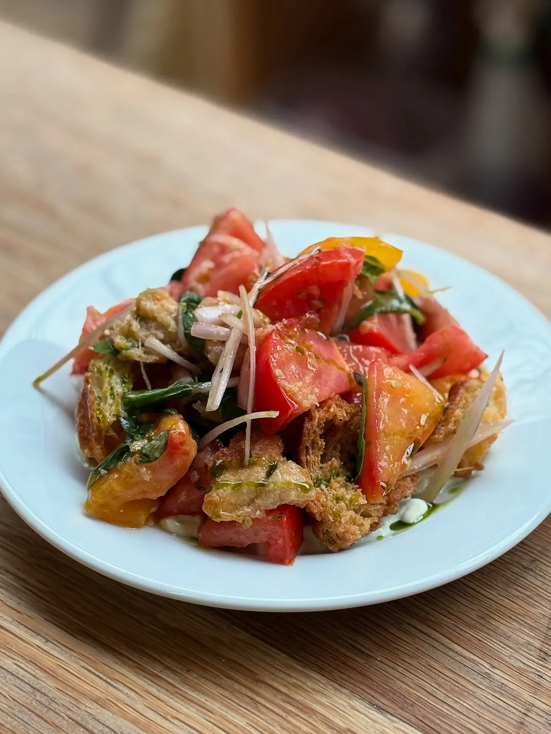 A close-up shot of a fresh tomato and bread salad, likely Panzanella, served on a white plate set against a wooden background. The salad features chunky red and yellow tomatoes, toasted bread pieces, thinly sliced red onions, and green herbs, all appearing lightly dressed.