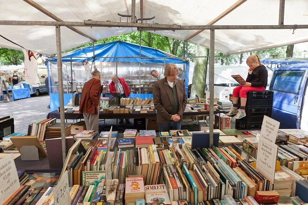 The image successfully portrays a vibrant book market scene. The composition is well-balanced, highlighting the array of books in the foreground and capturing individuals engrossed in browsing, including a charming detail of a child reading on a crate. Key elements such as the books and the central figures are sharp and in focus, contributing to the photo-realistic quality. The color accuracy is good, presenting natural tones. However, the lighting is somewhat flat and lacks dynamic range, which slightly reduces the overall visual impact, though it is evenly distributed.