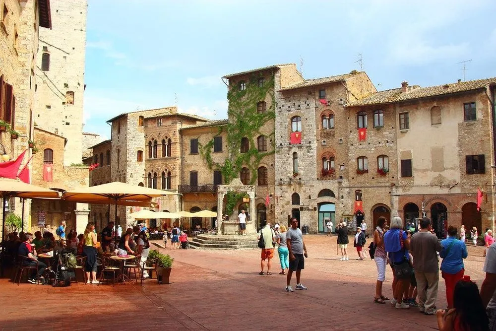 This image showcases a well-composed scene of a historic town square, featuring impressive architecture, a central fountain, and people. The natural lighting is good, illuminating the scene and bringing out the textures of the buildings. Colors appear accurate and vibrant, contributing to a strong sense of photo-realism. Key architectural elements, such as the buildings and the fountain, are sharp and in focus.