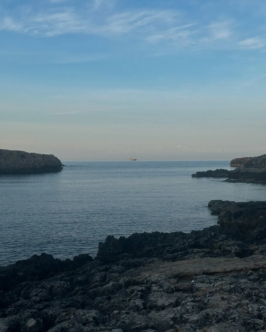 The image presents a serene coastal landscape featuring a bay framed by rocky formations under a clear sky. A small boat is visible in the distant calm waters.