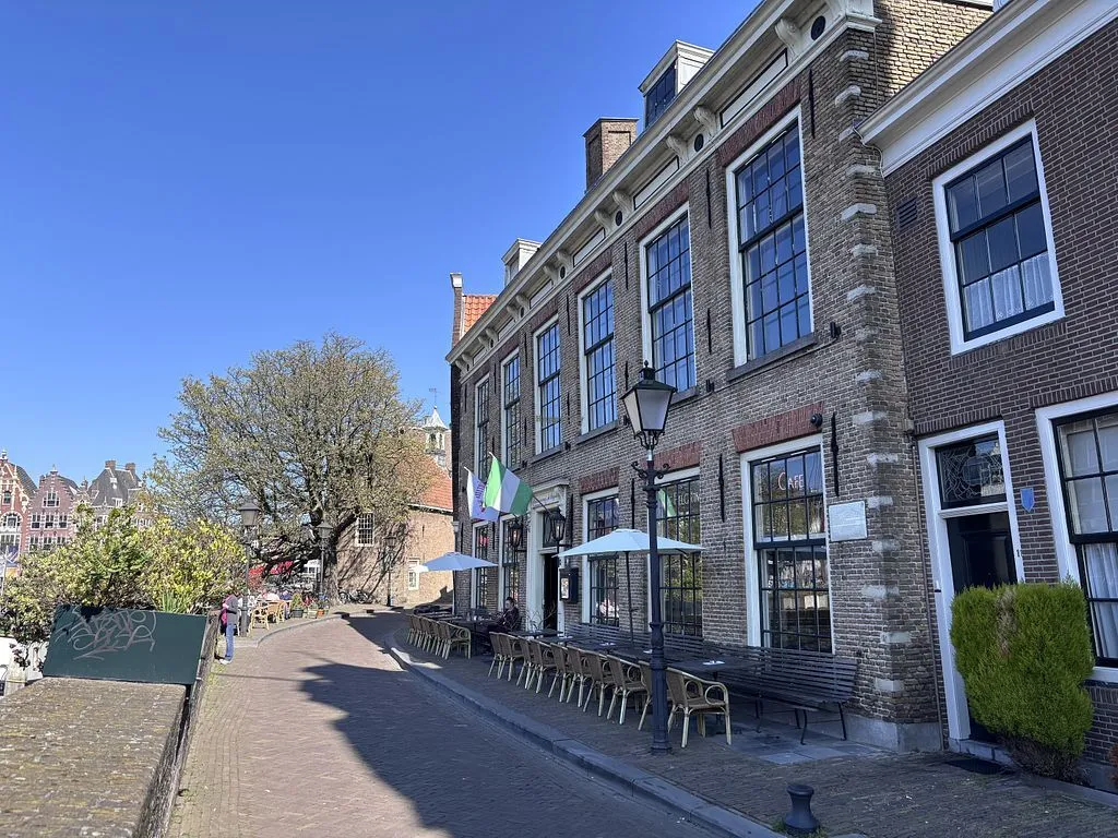 The image showcases a historic brick building housing a cafe with outdoor seating on a sunny day. The composition is well-balanced, and the natural light provides good illumination, though some shadows on the street are quite deep. The overall sharpness and color accuracy are excellent, contributing to a high level of photo-realism. The main architectural features of the building are in sharp focus.