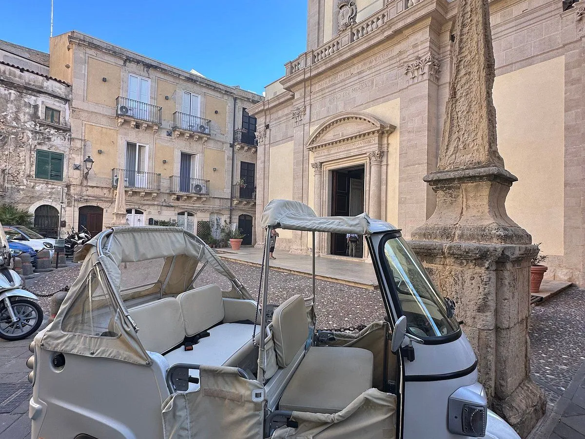 An editorial image showcasing a classic Ape Calessino vehicle parked in a historical European city square, with old stone buildings and a clear blue sky in the background.